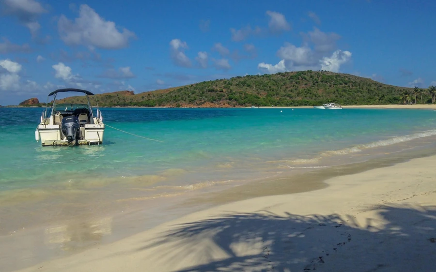 A small white boat anchored off a long pale sandbar with green hills behind