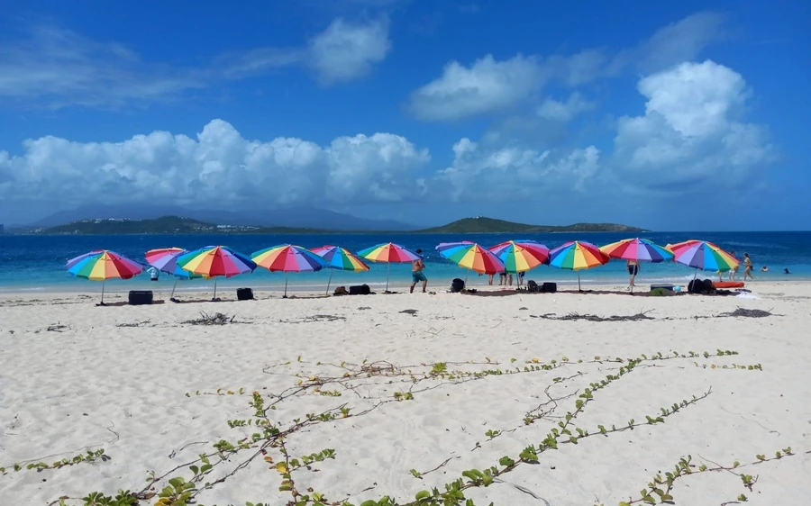 A row of rainbow-coloured beach umbrellas on white sand under a blue sky