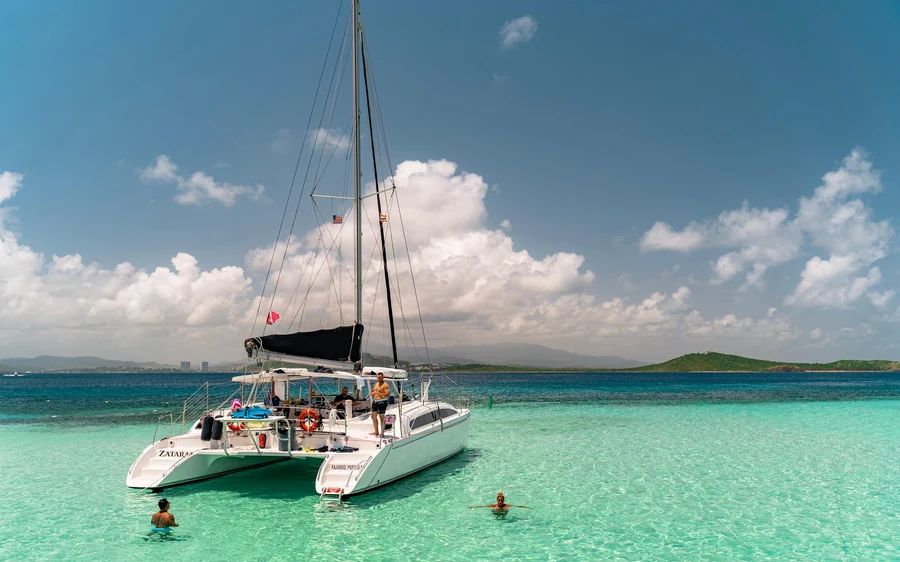 A sailing catamaran anchored off a green island with people in the water beside it