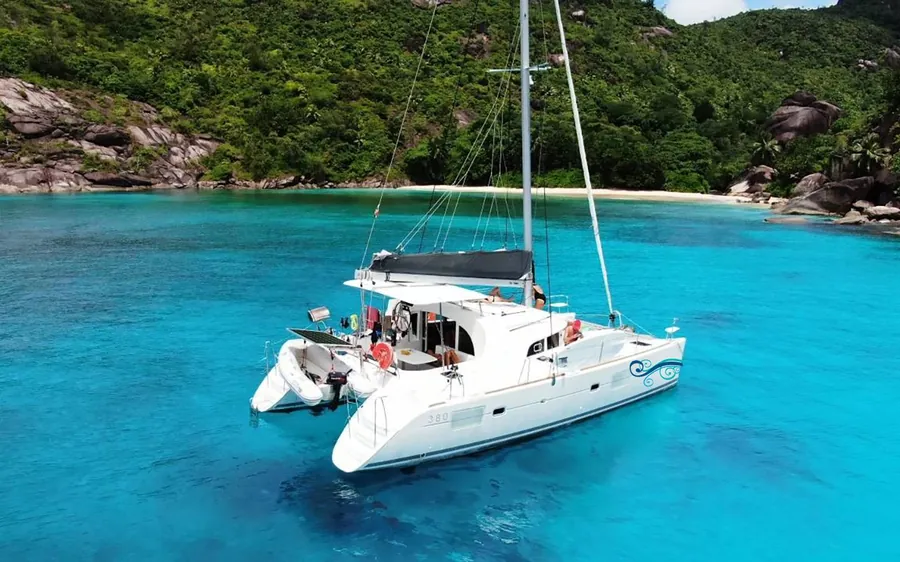 A white sailing catamaran at anchor in turquoise water with green hills behind
